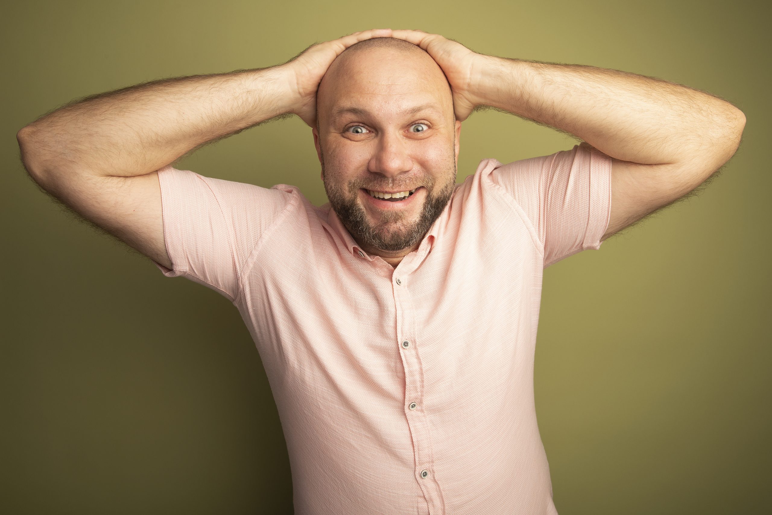 joyful middle-aged bald man wearing pink t-shirt holding hands on behind head isolated on olive green background